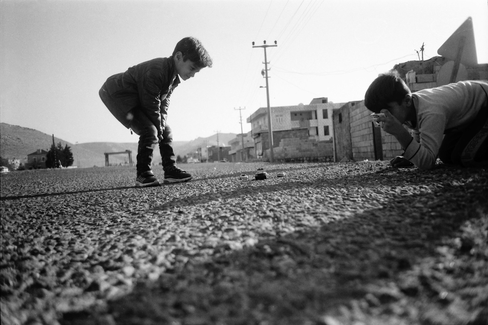 A young boy crouches in the street while another boy takes photos of objects arranged in front of him 
