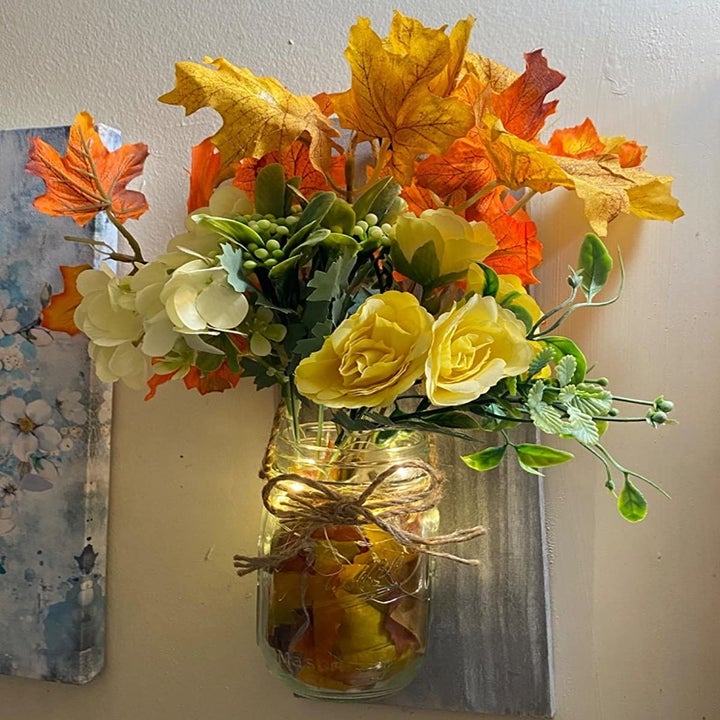 A clear mason jar with fairy lights in the bottom and fake greenery and yellow roses at the top. Orange fall leaves have also been added.