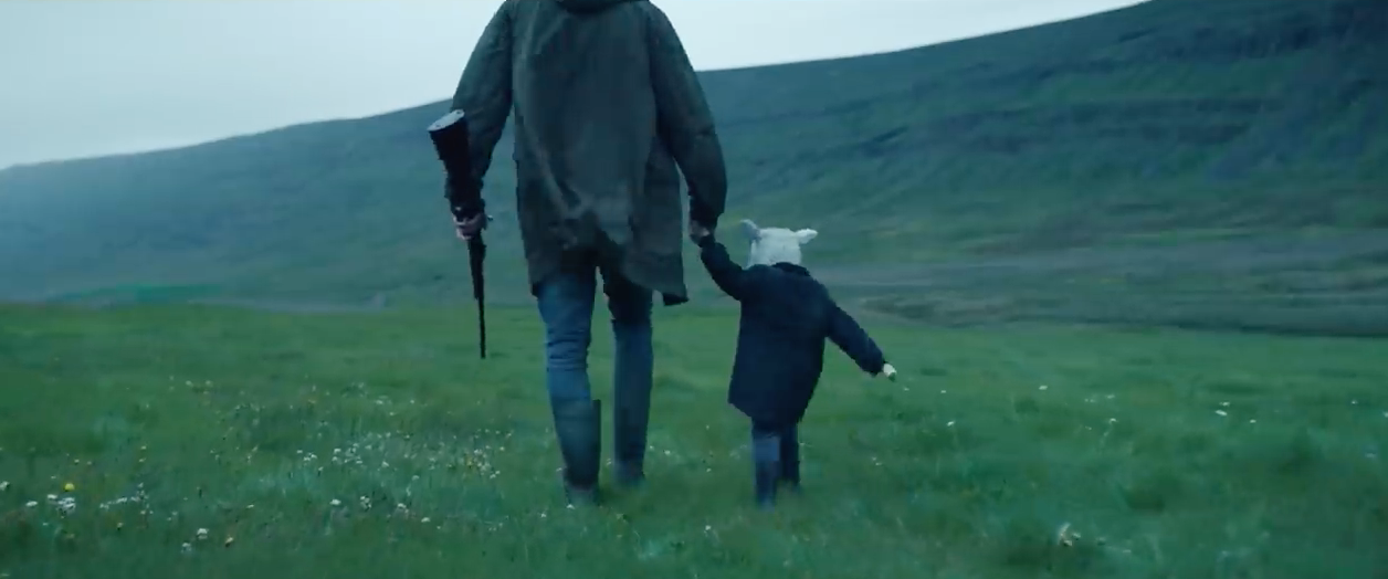A man holding the hand of a half sheep half child as they walk through a field