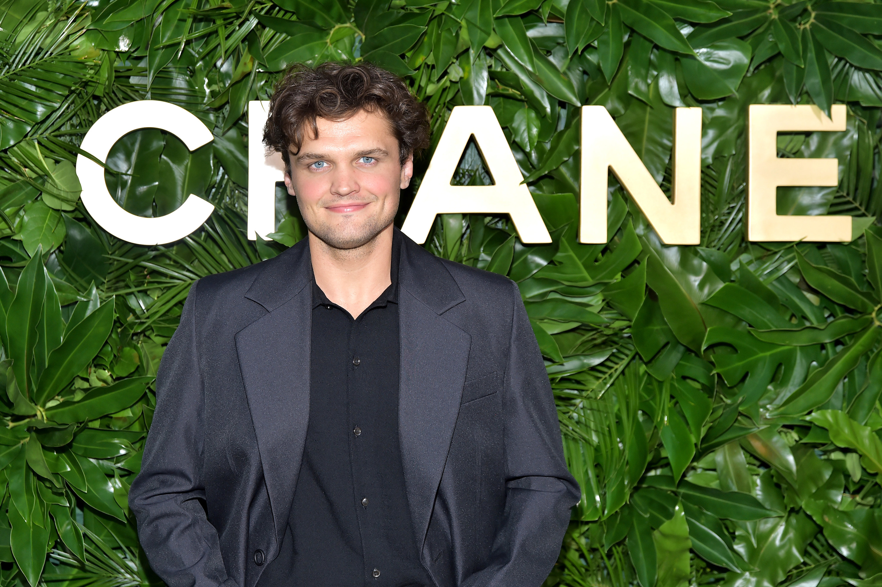 Smiling Ray in a shirt and suit jacket stands behind a leafy backdrop with &quot;Chanel&quot; logo