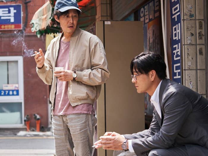 Gi-hun stands next to a seated Sang-woo; they both smoke
