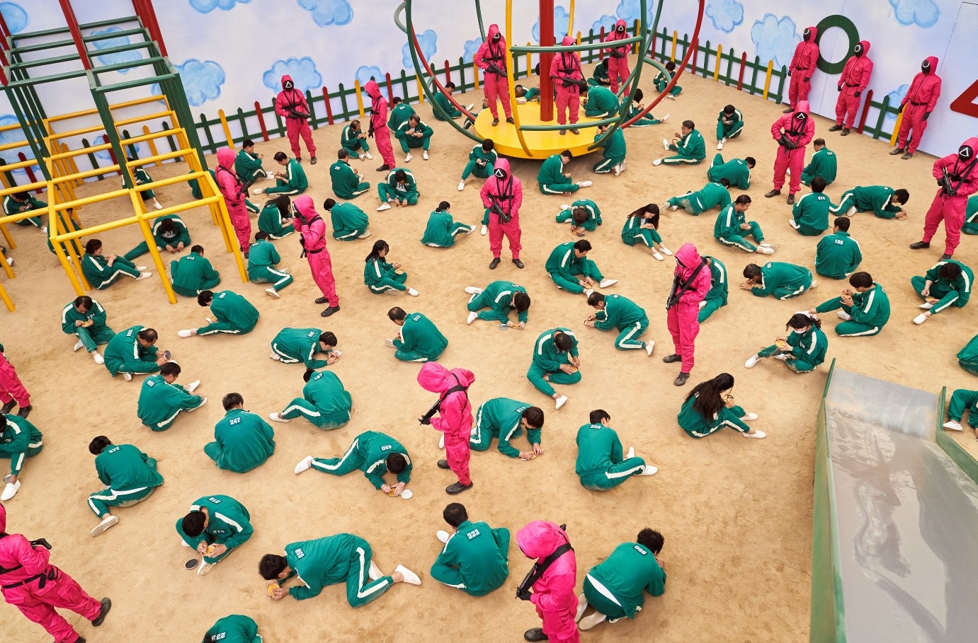 The contestants kneel on the ground in a huge playground set with the guards watching over them