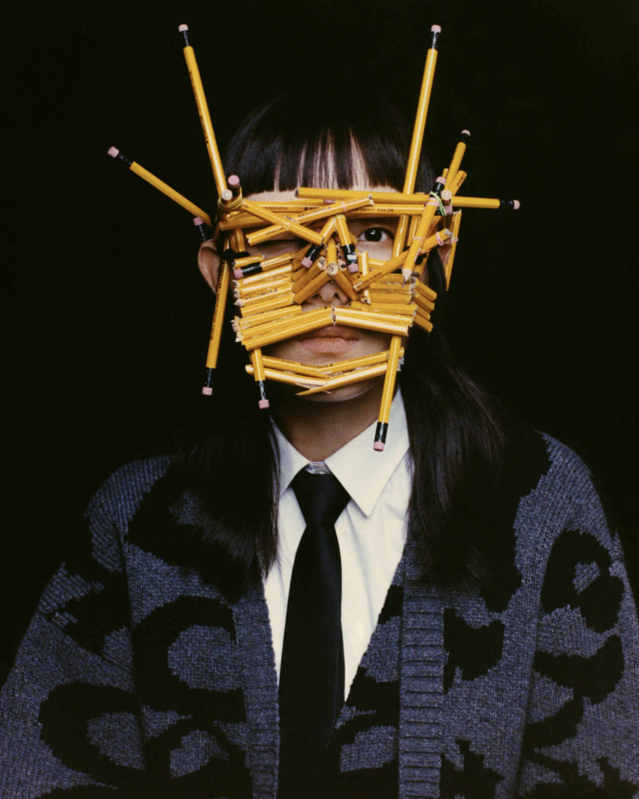 A person in a sweater looks at the camera, with their face covered by numerous pencils bundled together in several directions