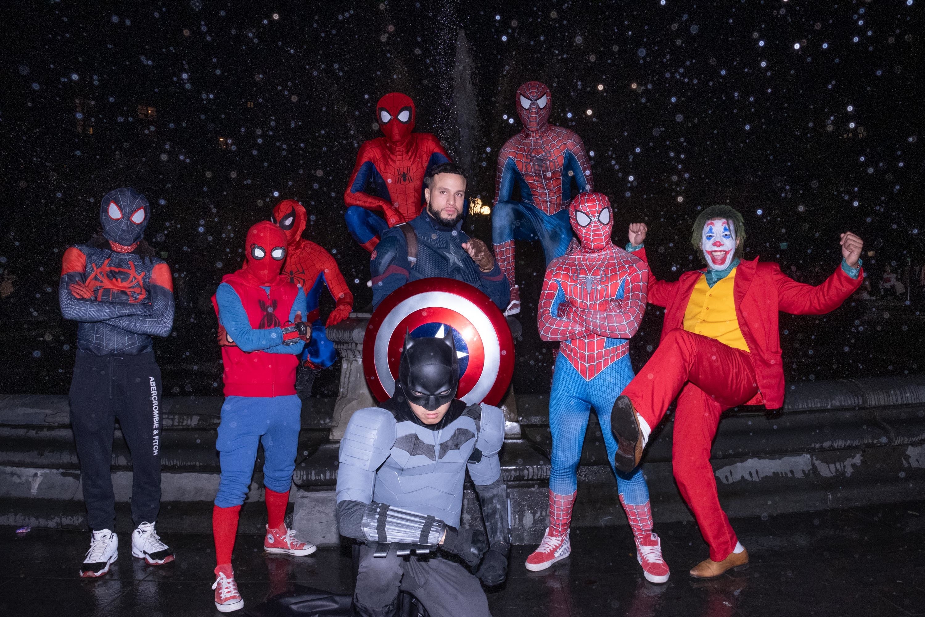 Six people dressed as spiderman, one batman, a captain america and a joker in front of a fountain