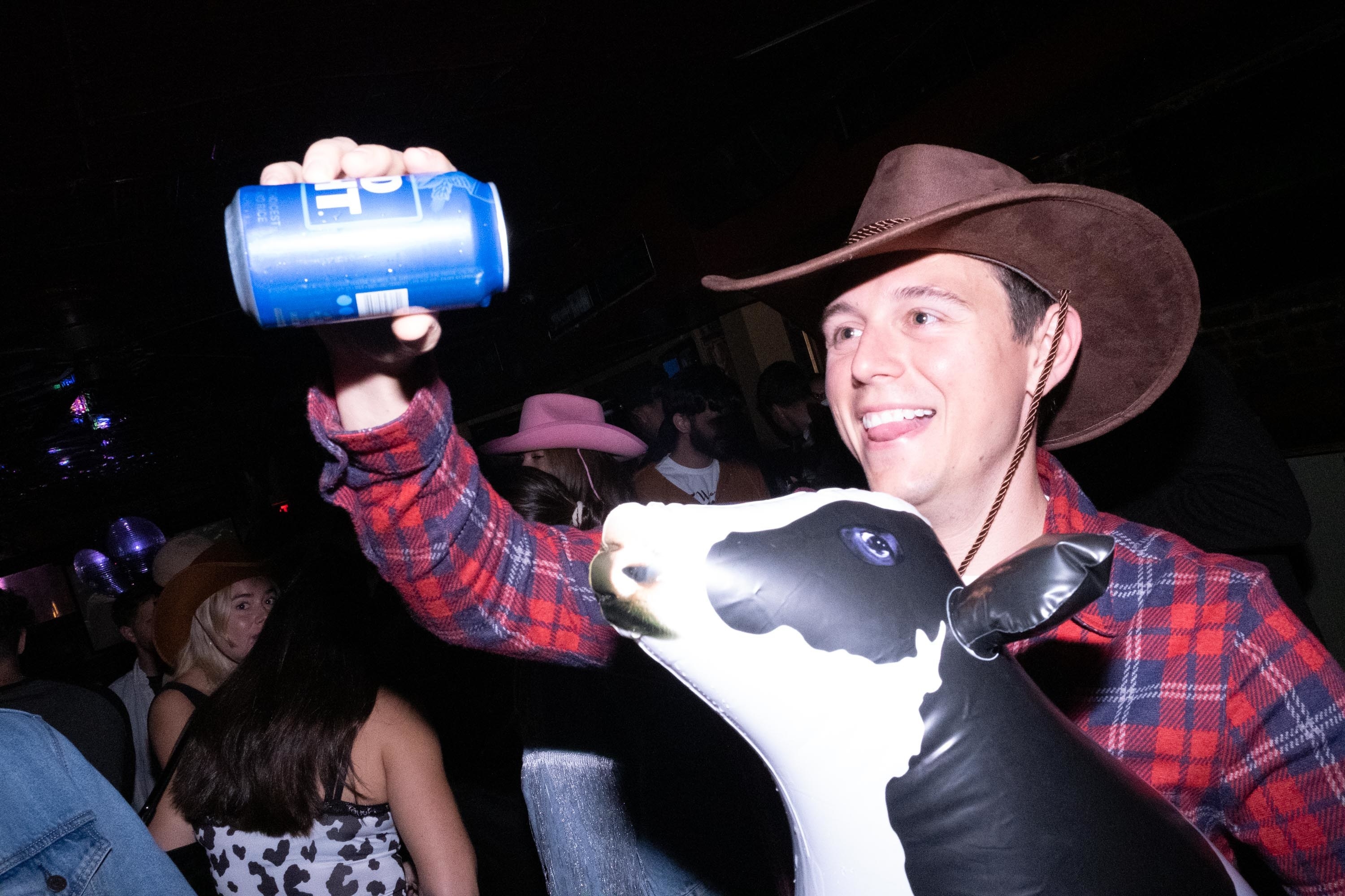 A man in a cowboy hat pretends to pour a beer into the mouth of an inflatable cow