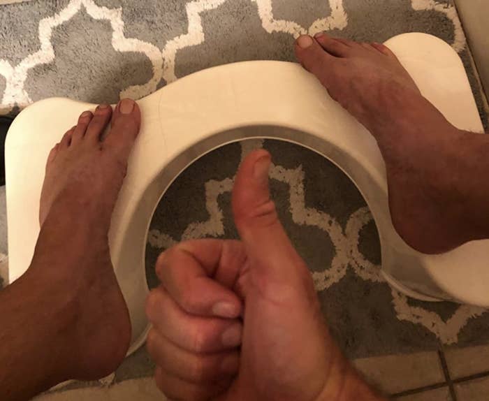 A customer review photo of them holding up a thumbs up while their feet are on the squatty potty which is placed next to their toilet