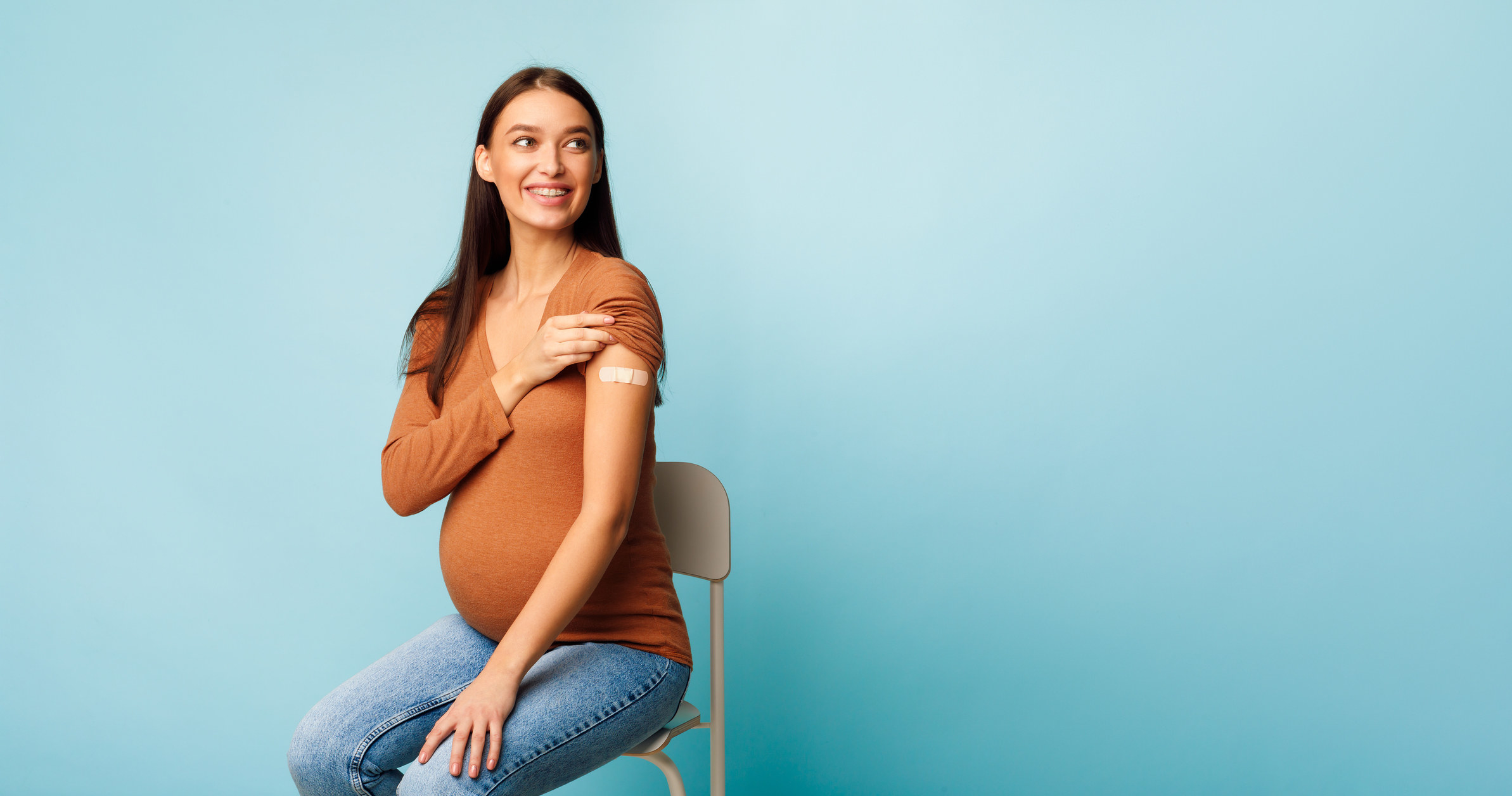 A pregnant woman sits on a chair. She is proudly displaying a plaster on the top of her arm where she has been vaccinated.