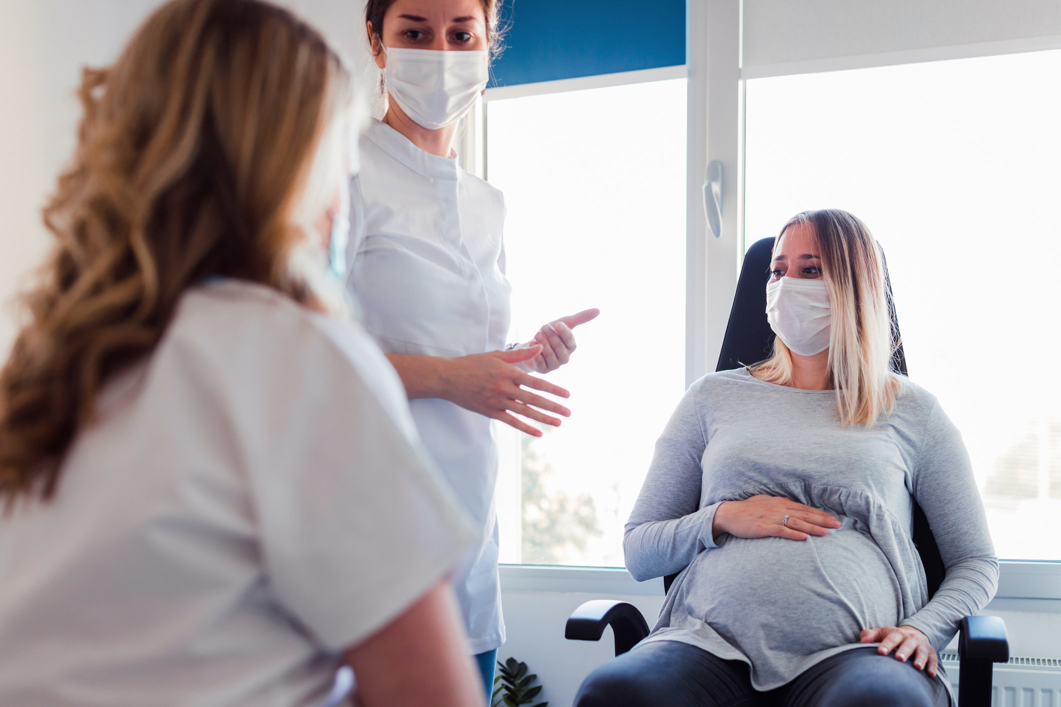A pregnant woman sits with a mask on in a doctor&#x27;s office while two people watch over her