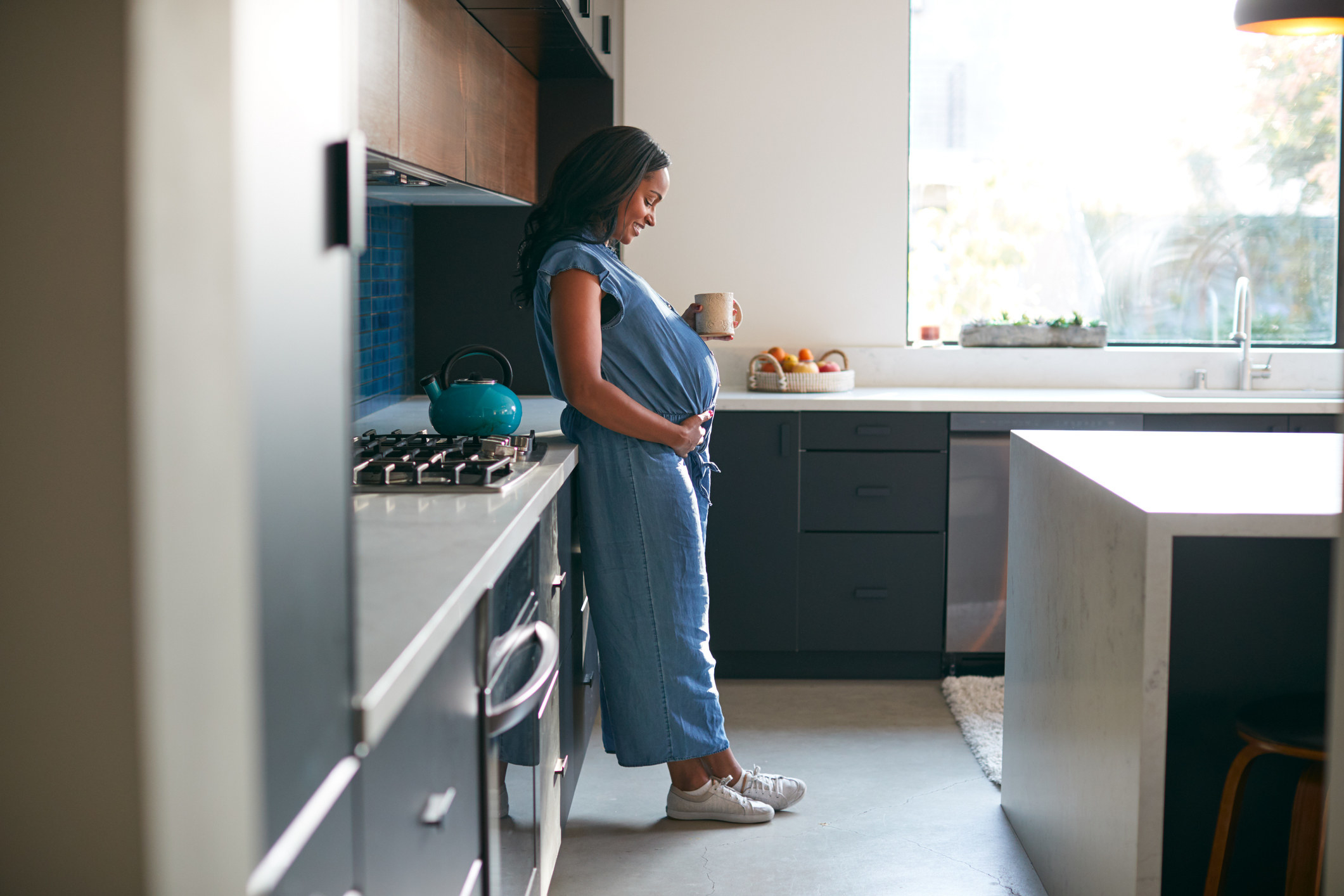 A pregnant woman stands in her kitchen enjoying a cup of tea and holding her bump