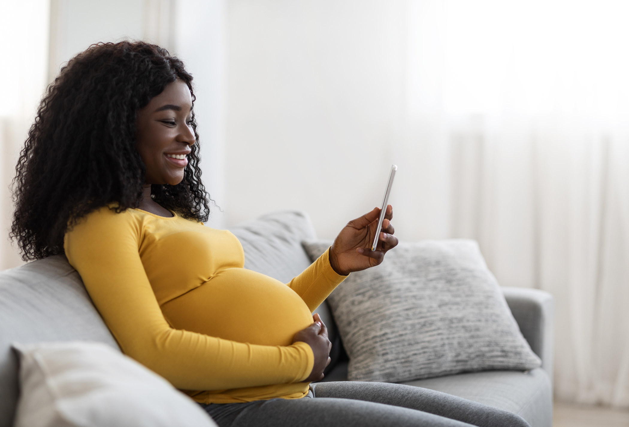 A pregnant woman reclines on her sofa while looking at her phone and smiling