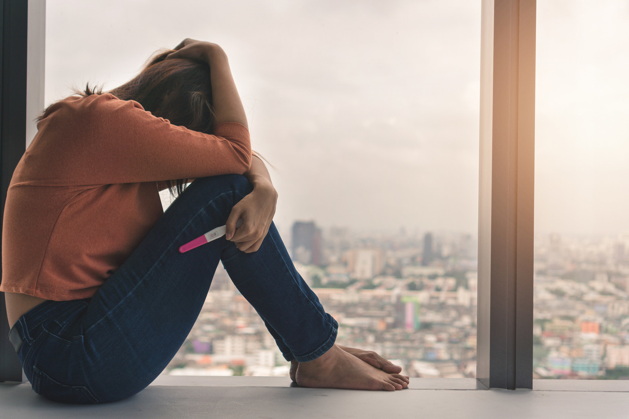 A woman sitting on a window sill with her head down, holding a pregnancy test