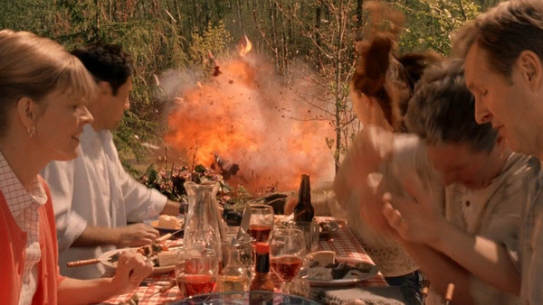 A group of people all sit around a table eating at a picnic, and in the middle of the shot, there is a sudden fire explosion where the grill was