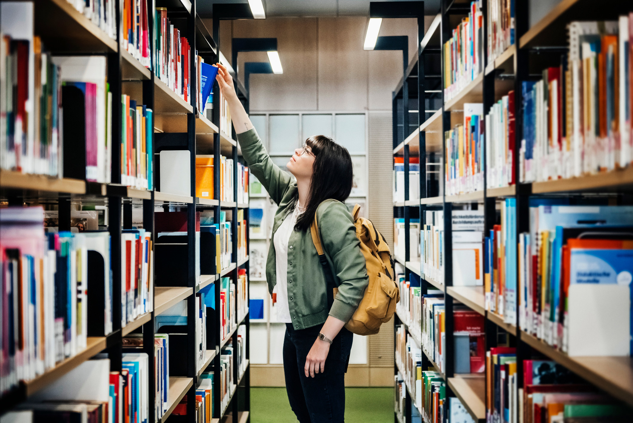 A young student reaching for a book from the top shelf in a public library
