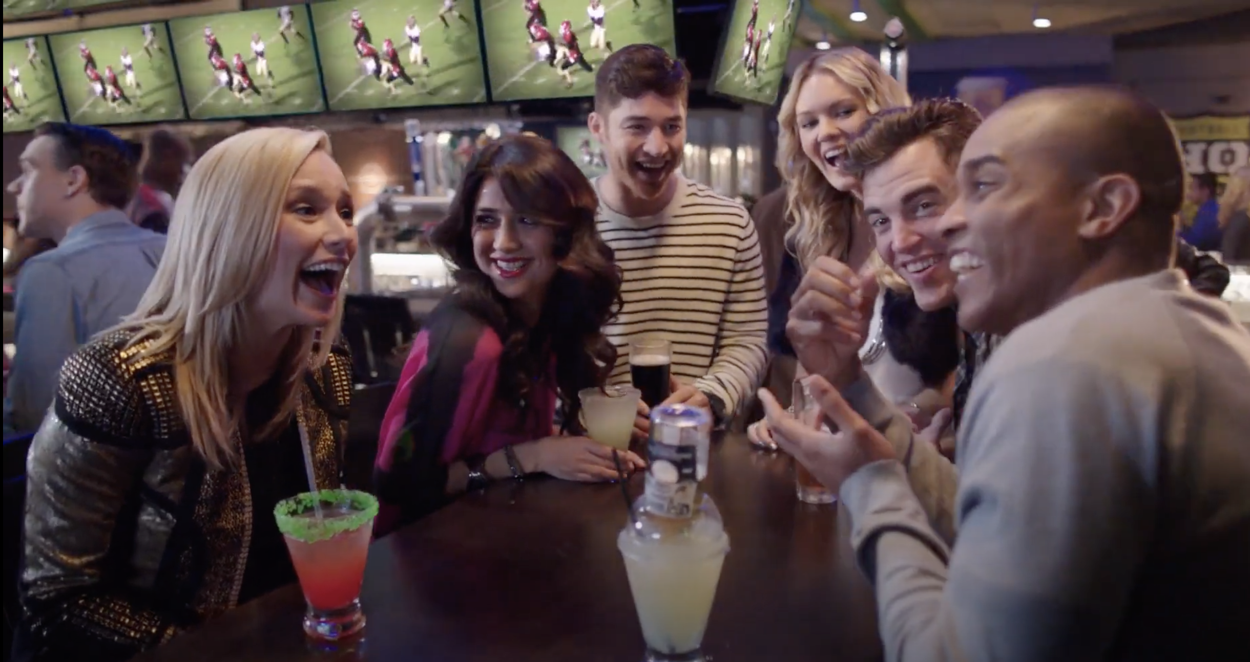 A group of friends smile and laugh in a bar with football on the TVs in the background