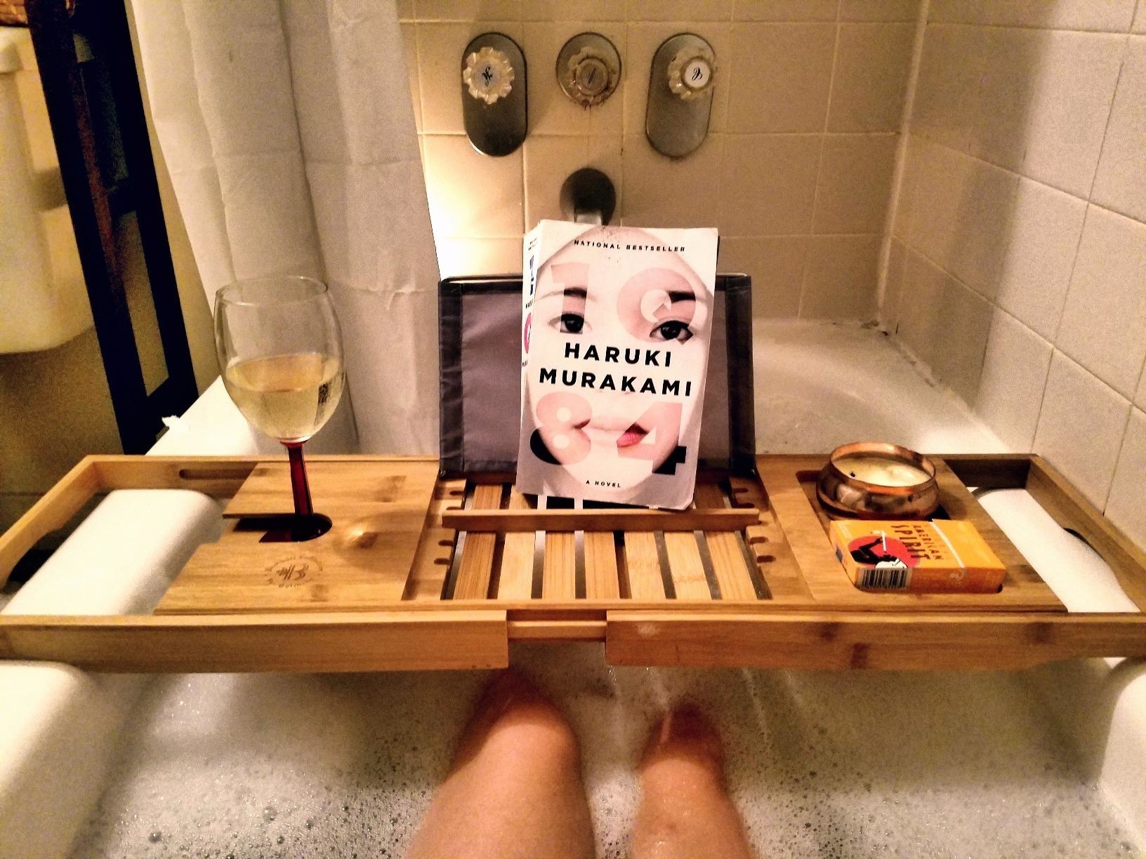 Reviewer's light wood bath caddy with a glass of white wine, a book, and candle over their bath tub