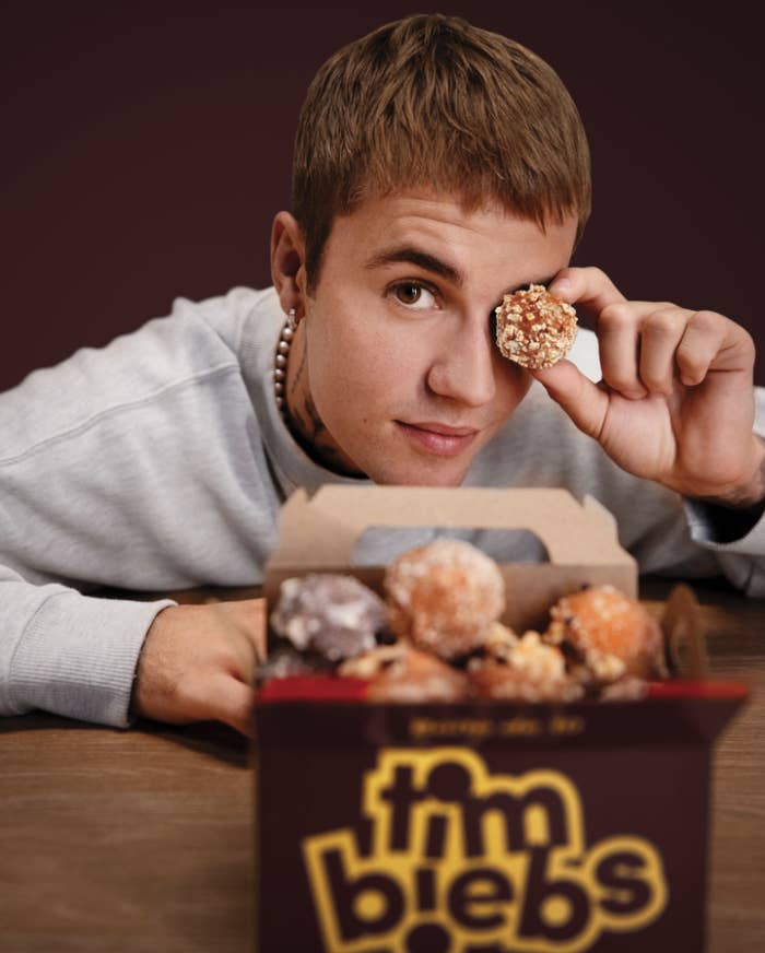 A photo of Justin Bieber holding a timbit up to his eye with a box of Timbits in front of him.