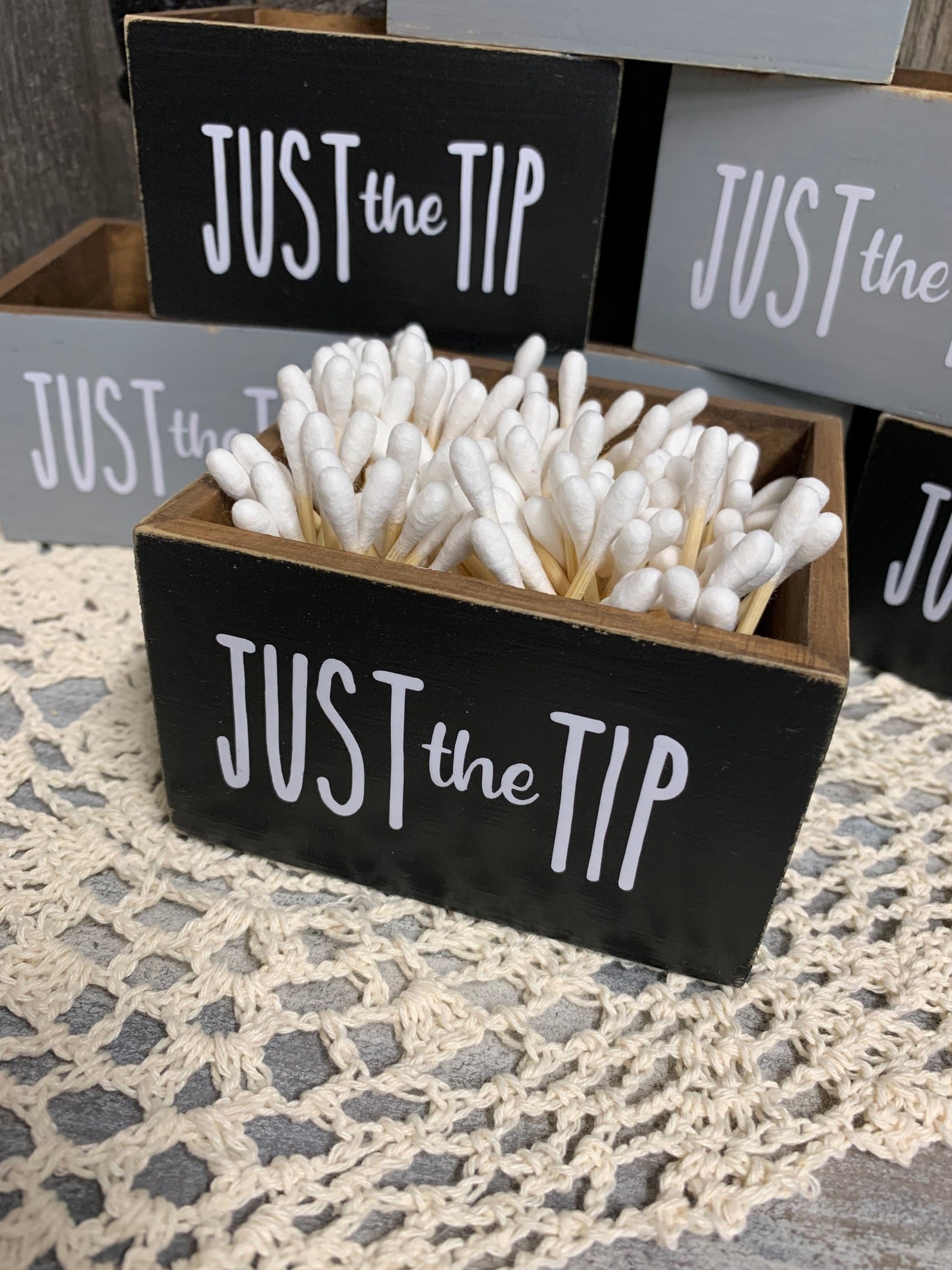The black &quot;Just The Tip&quot; cotton swab holder in front of a stack of black and gray cotton swab holders