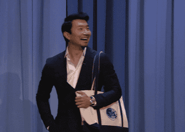 simu liu arriving at the tonight show starring jimmy fallon carrying a bag and waving with a big smile on his face