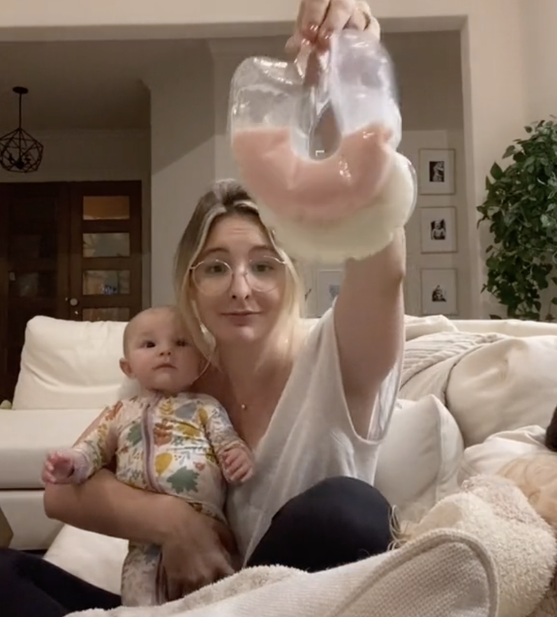 Jo holding her baby in one arm and two bags of her breastmilk, one white in color and the other pink, in the other arm
