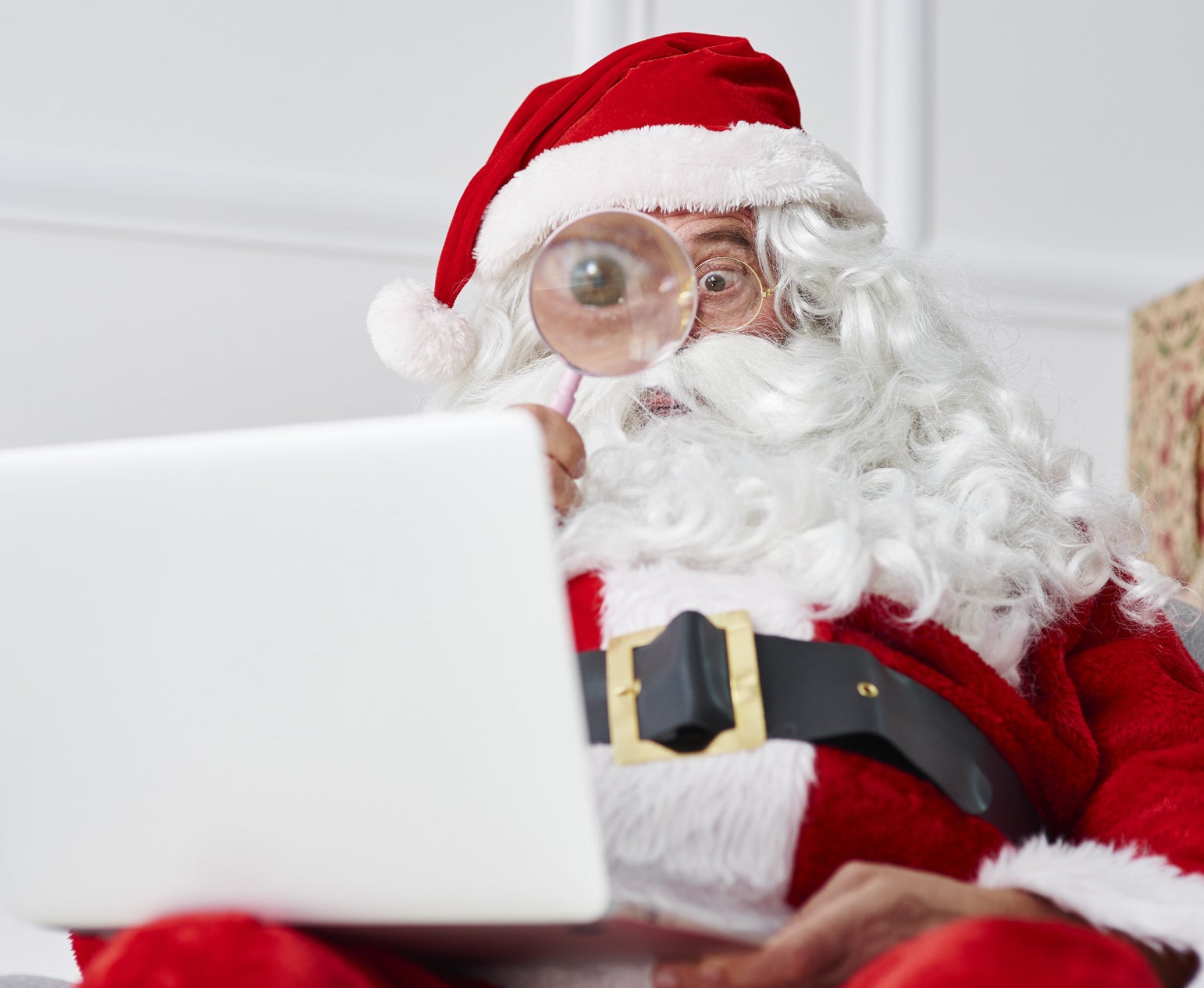 A photo of Santa sitting down with a laptop on his knee holding a magnifying glass up to his eye.