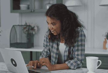 A GIF of a woman standing in her kitchen looking at her laptop and getting excited.