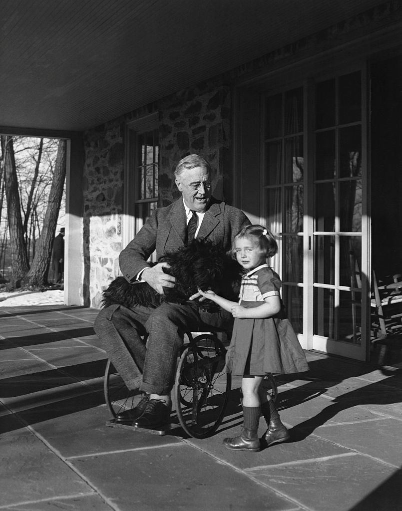 Franklin Roosevelt in a wheelchair holds his Scotch terrier, Fala, on his lap as he talks to Ruthie Bie, the daughter of the Hyde Park caretaker