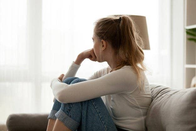 A girl sits on the couch with her legs pulled up to her chest with one arm wrapped around her knees and the other pressed into a fist against her mouth as she turns to look out the window