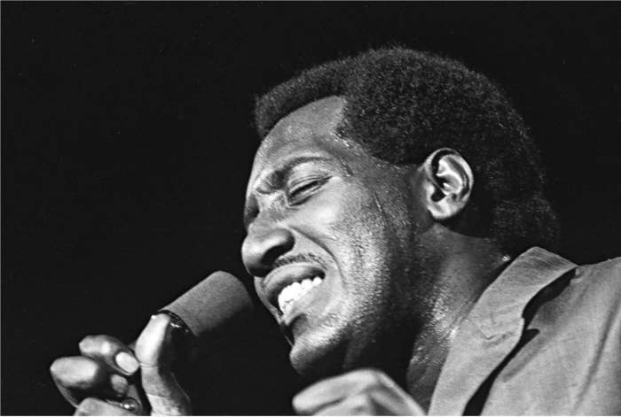 Otis Redding on stage, in a light colored, double breasted suit, mike in right hand, profile, spotlight above