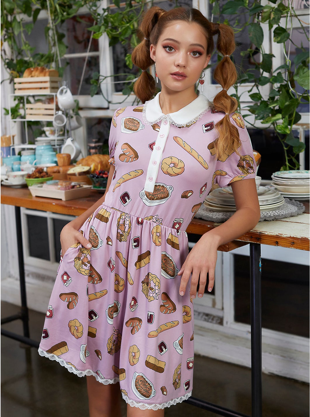 Model in short sleeve pink peter pan collar dress with white lace trim and print of various foods and baked goods from the movie