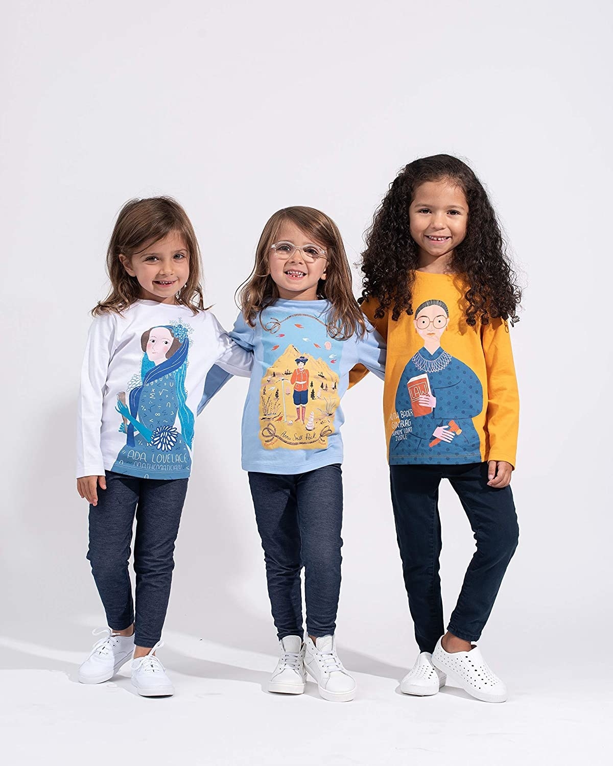 Three girl models wearing long-sleeve Piccolina shirts each with a powerful female graphic on the front: Ada Lovelace, Annie Smith Pock, and Ruth Bader Ginsburg