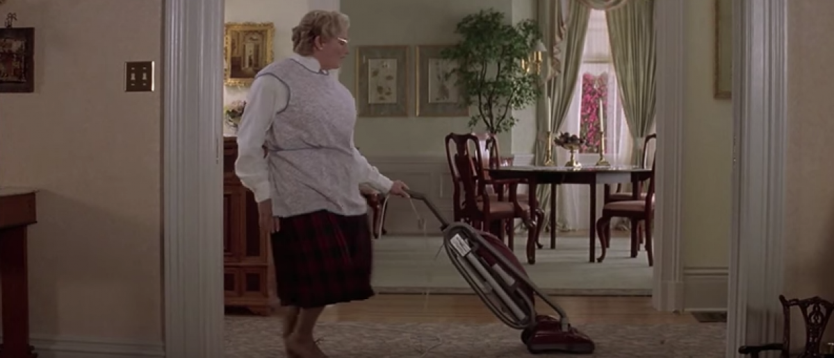 A man disguised as an older woman vacuuming in the living room.