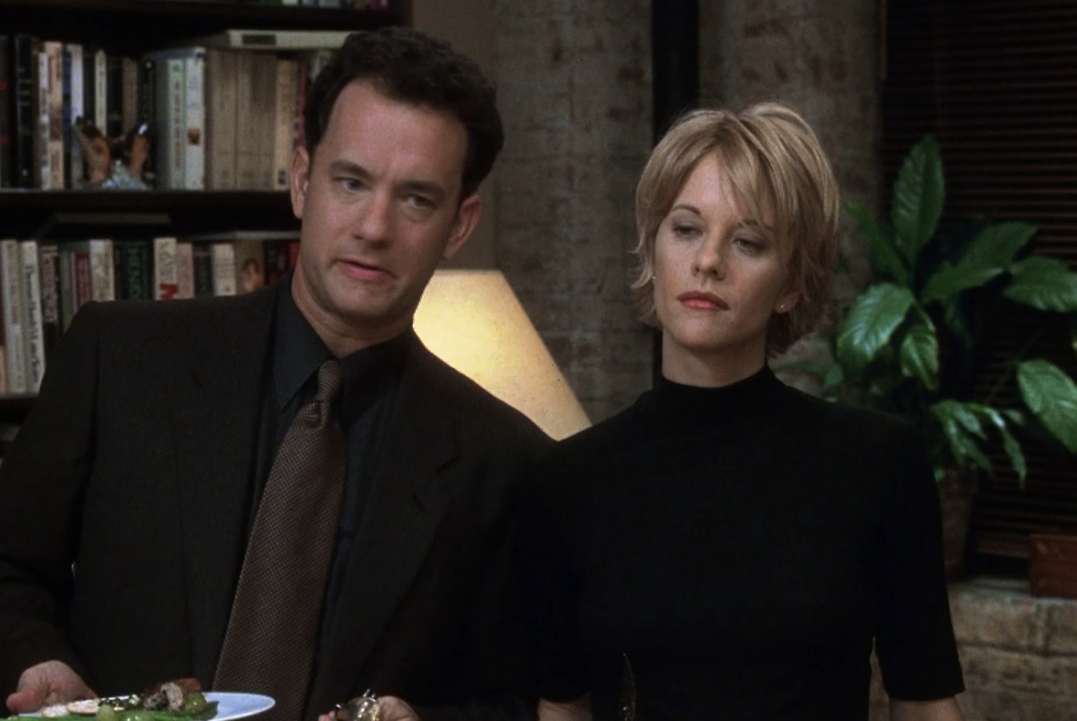 Actors Tom Hanks and Meg Ryan stand next to each other but look ahead. Hanks has a plate of food in his hand.