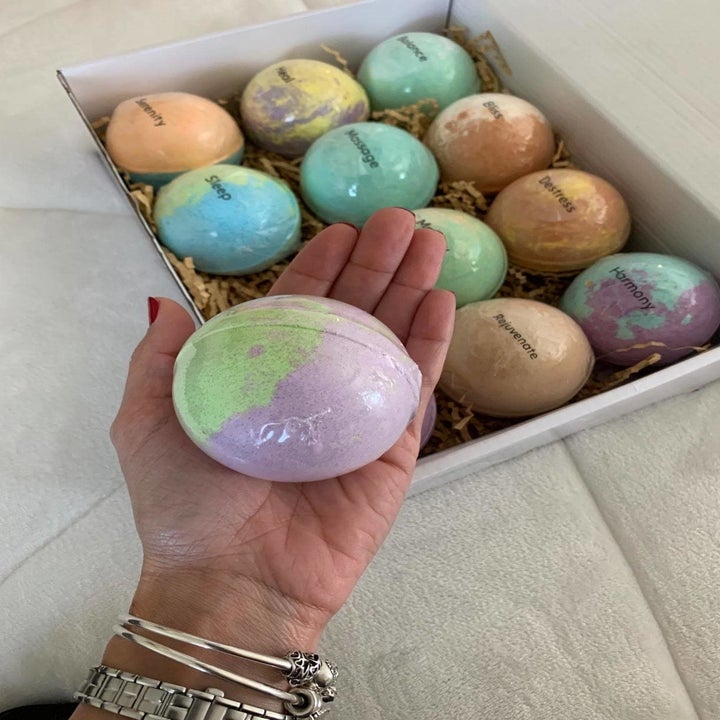 A reviewer's hand with a large bath bomb, and a box with the colorful bath bombs inside