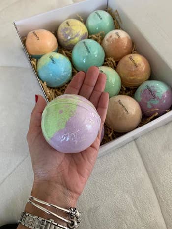 A reviewer's hand with a large bath bomb, and a box with the colorful bath bombs inside