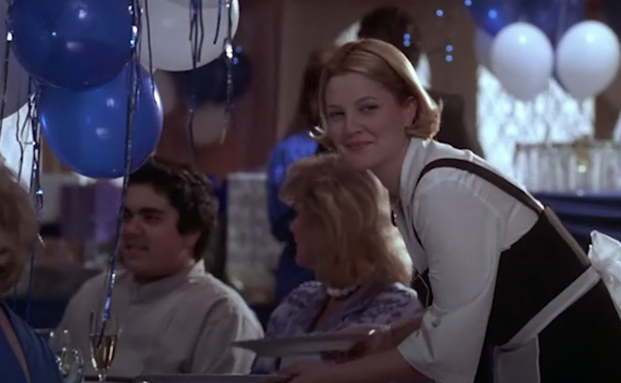 Actor Drew Barrymore is dressed as a server and waits on a table. Blue and white balloons are in the background.