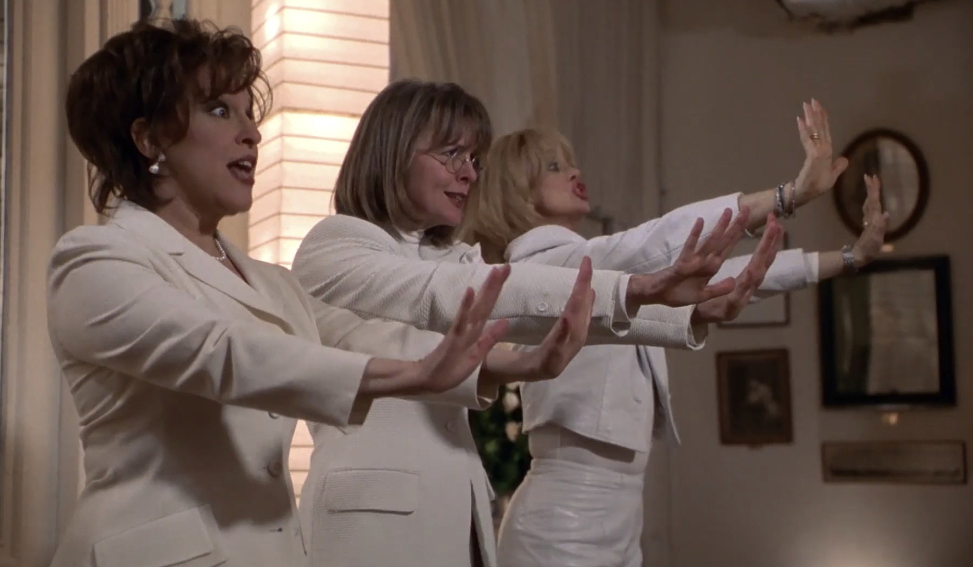 Actors Bette Midler, Diane Keaton, and Goldie Hawn wear all-white outfits. They have their hands up before them as they sing.