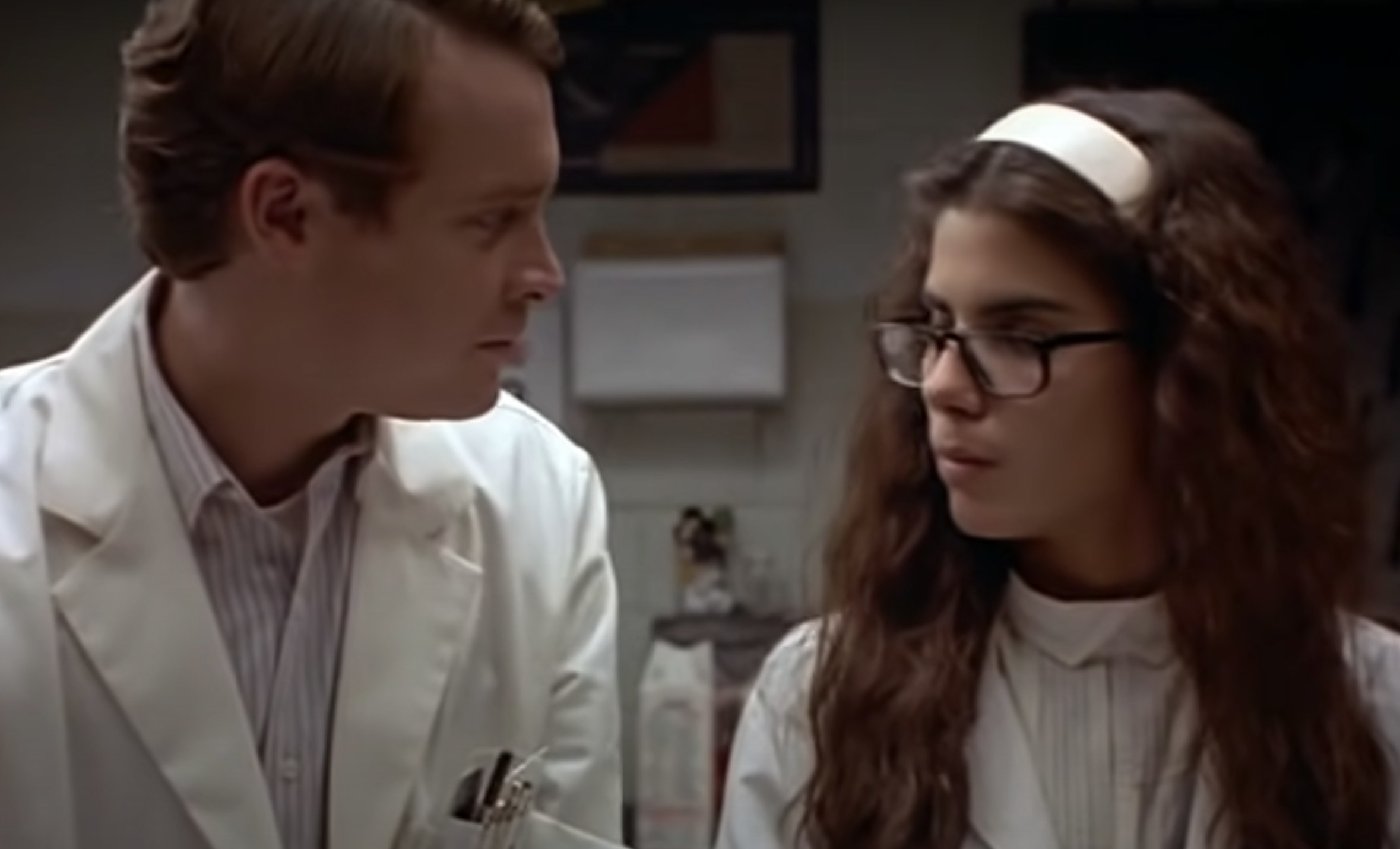 Actors Tate Donovan and  Sandra Bullock wear lab coats and look at each other as they speak.