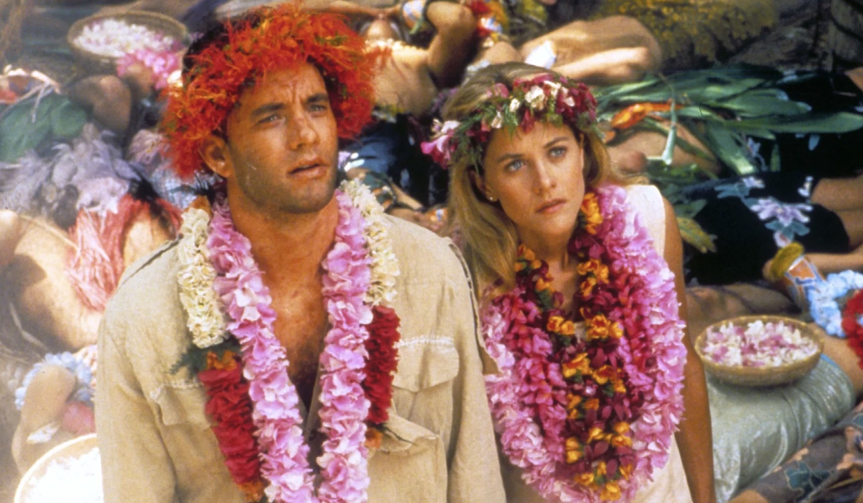 Actors Tom Hanks and Meg Ryan stand next to each other looking dumbfounded. They wear necklace and crowns made of tropical flowers.