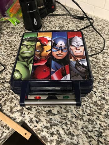 A reviewer showing the square waffle iron with Hulk, Iron Man, Captain America, and Thor pictured on top