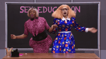 two drag queens in front of a sex education chalk board