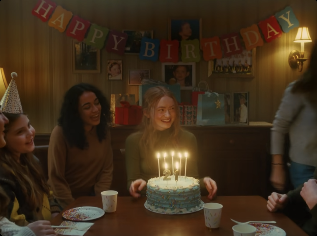 Her sitting at a table in front of her birthday cake and surrounded by friends