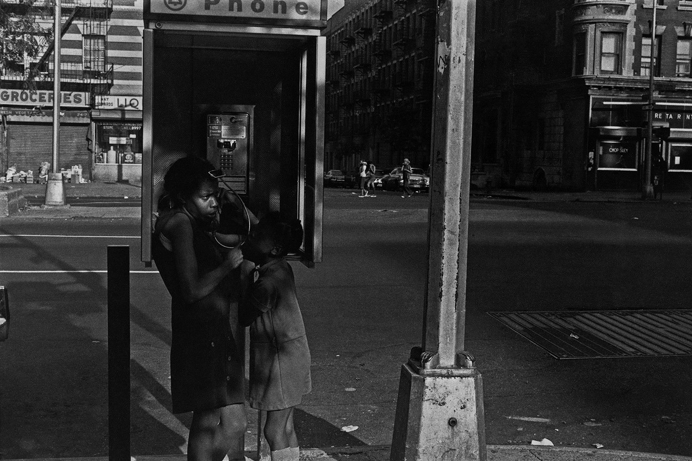 A mother and child at a payphone on an empty street, lamp post in the foreground 