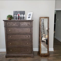 the mirrored jewelry armoire next to a brown dresser with paintings on top of it