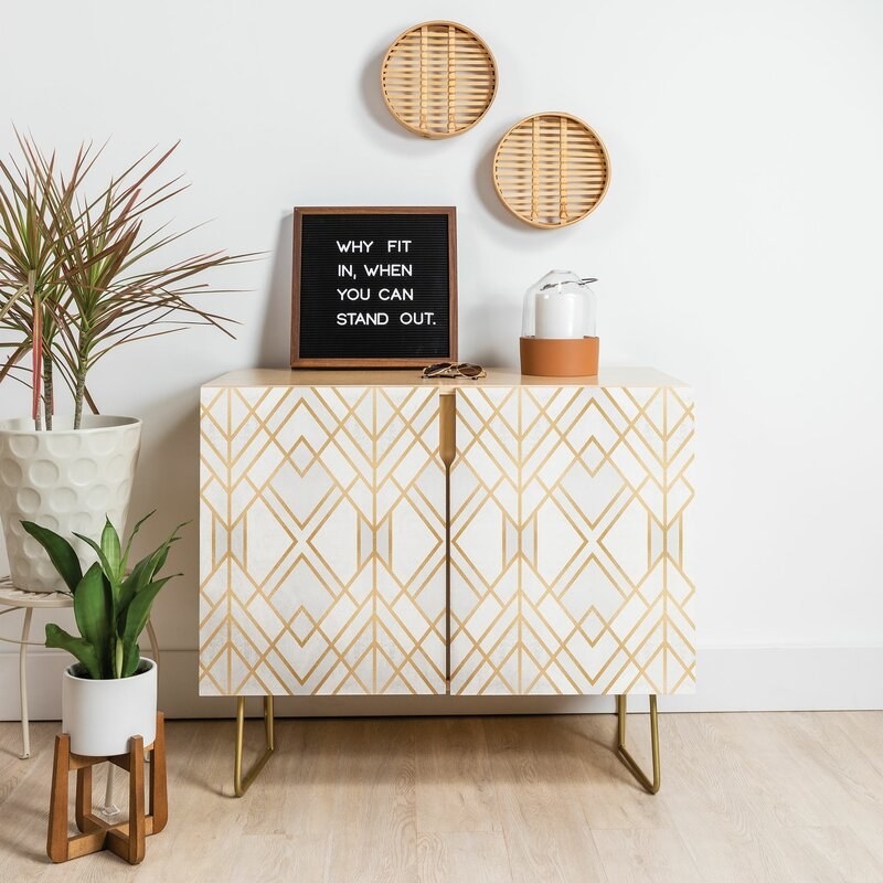 the cabinet with a white and gold geometric pattern, a plant in front of it, a candle on top and a sign that says &quot;why fit in when you can stand out?&quot;