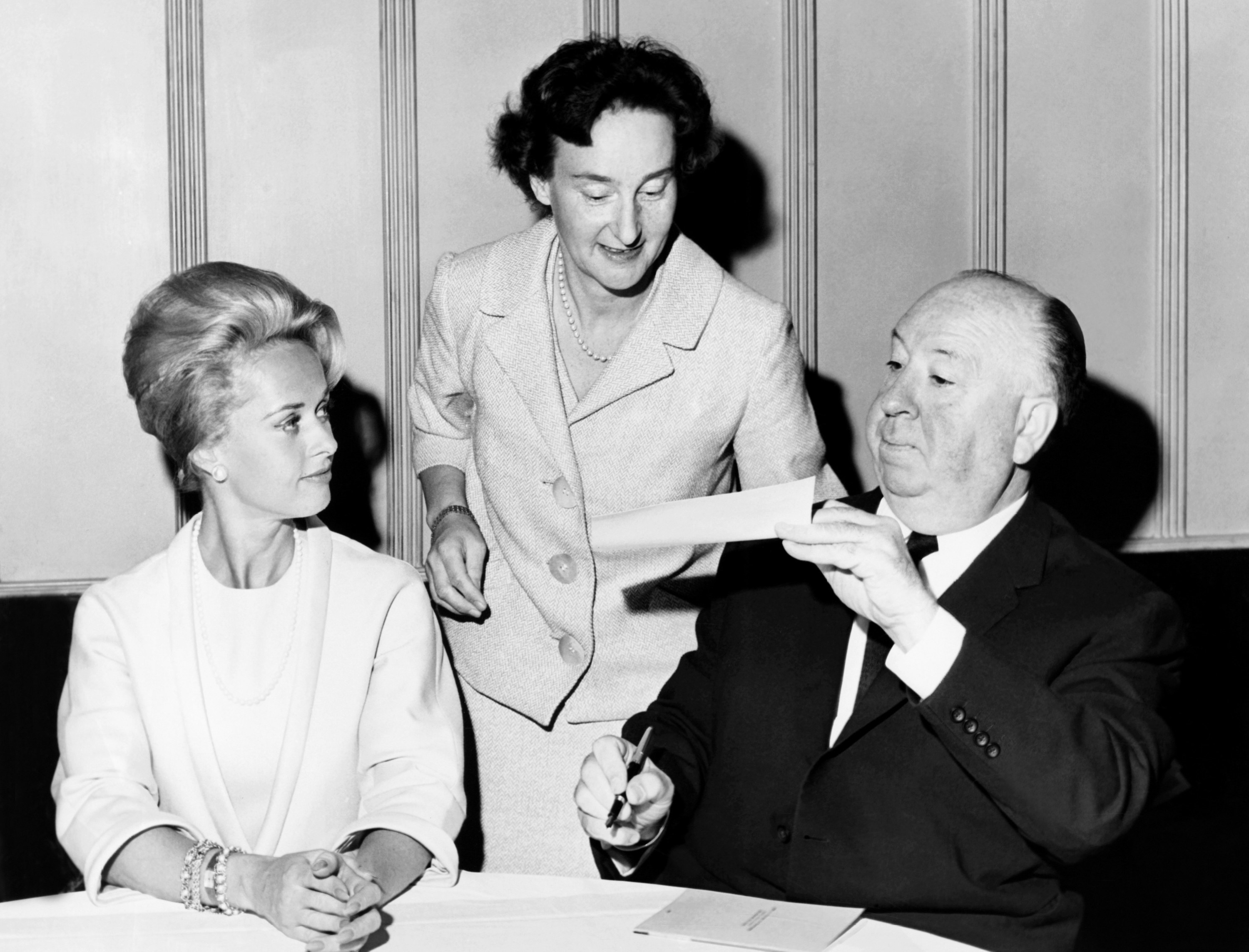 Hitchcock hands someone a piece of paper with Hedren sitting next to him
