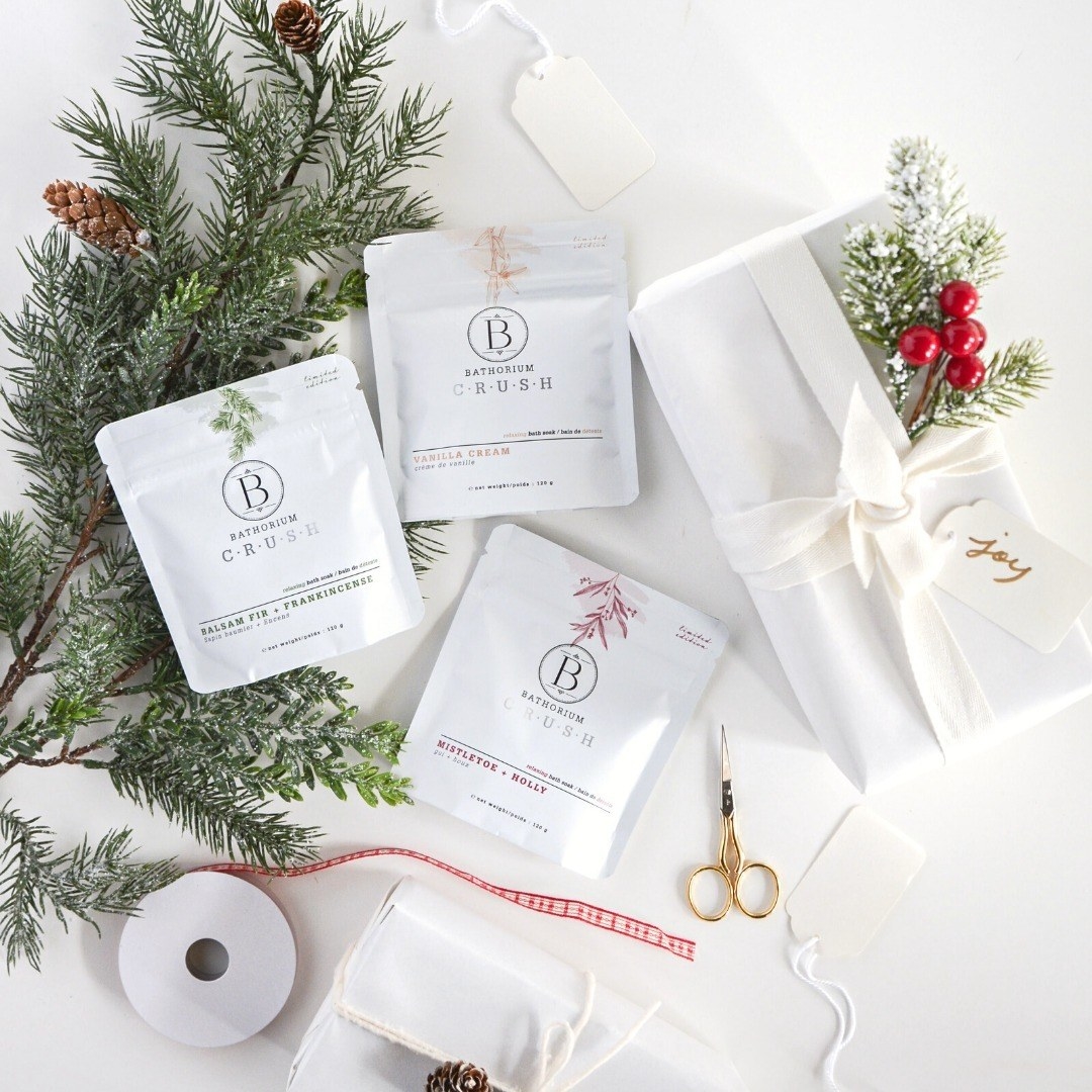 a trio of limited edition bath salts in holiday scents surrounded by wrapping paper and fresh pine boughs