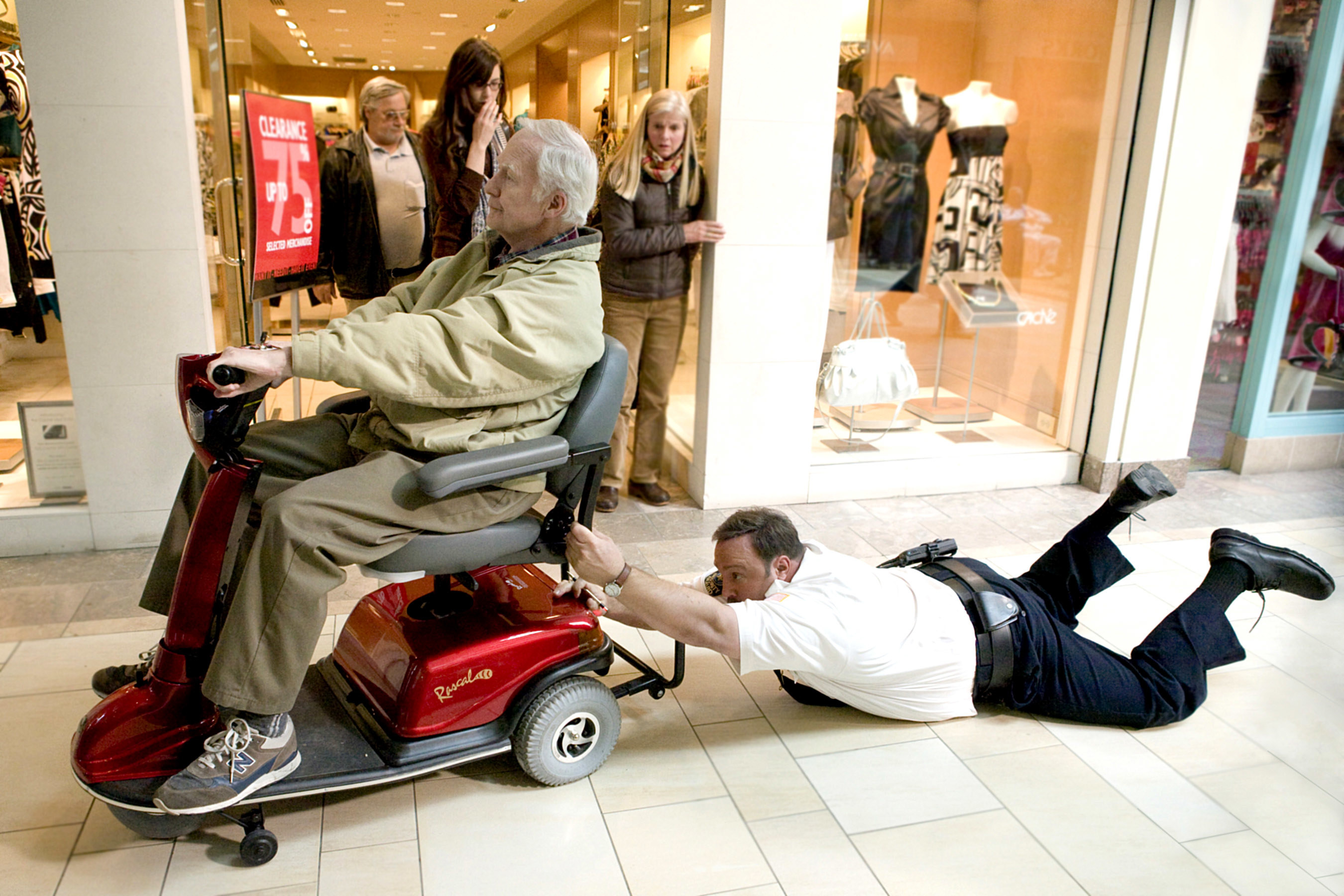 Paul Blart holding onto a man in a motor scooter and being dragged across the mall floor