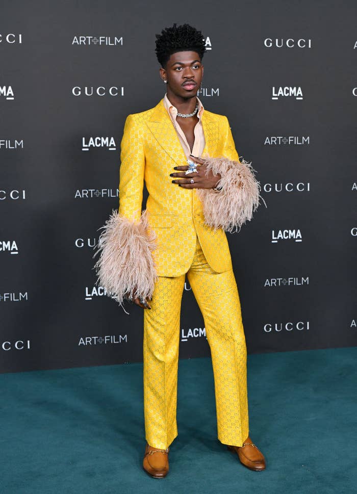 Lil Nas X at a red carpet event rocking a brightly-colored textured suit with feathers on the ends of the sleeve