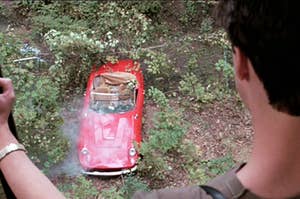 A man's back is facing away from the camera. He looks down at a red car that has crashed in the forest and looks totaled Credit: Cbs Photo Archive / CBS via Getty Images