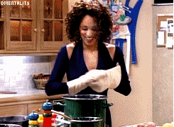 A woman happily drops an ingredient into a pot of water on the stove